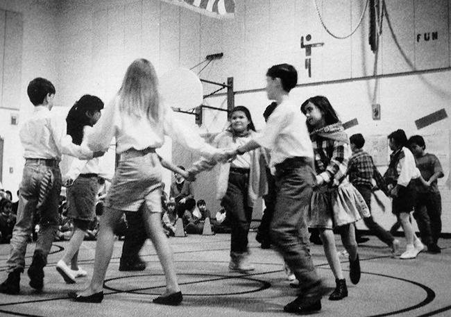 Black and white yearbook photograph of students dancing in the gymnasium. Small groups of students are holding hands and moving in circles.