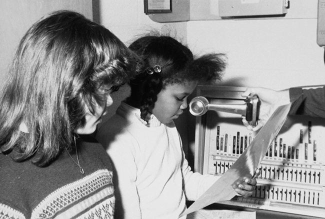 Black and white yearbook photograph of two students reading the morning announcements into the school-wide address intercom system’s microphone.