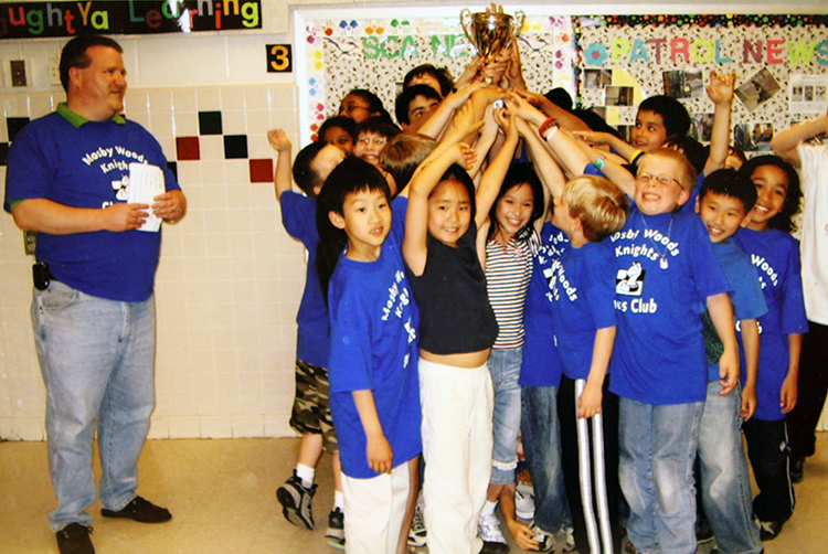 A group of students wearing blue Mosby Woods Knights Chess Club t-shirts proudly hold up a gold trophy cup.