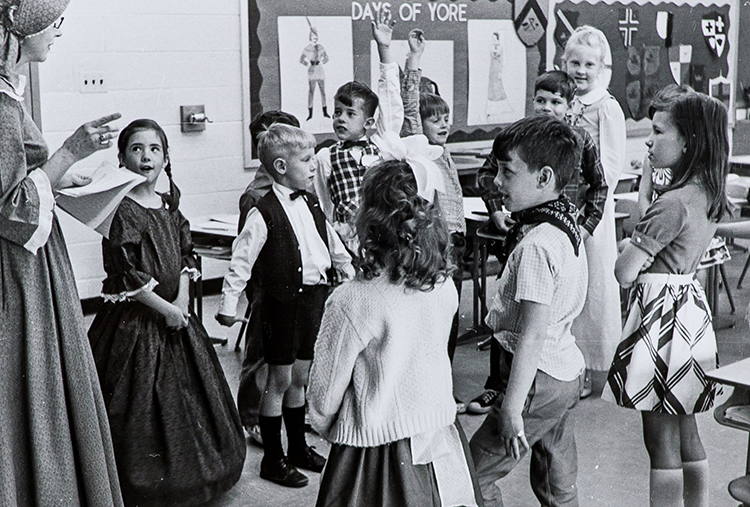 Black and white photograph of a small group of students participating in a play. They are dressed in old fashioned clothing from the 19th century. 